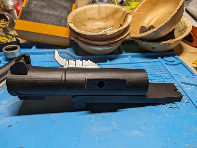 Black resin barrel and receiver section on a blue cutting mat with a silver printed trigger part placed behind it and a rectangular cutout in the barrel