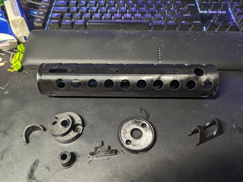 Black resin barrel shroud with a row of ten cooling holes lying on a desk mat, surrounded by small printed rings, clips and a muzzle piece
