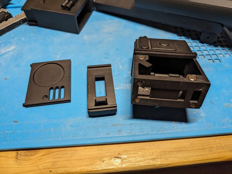Three black resin parts on a blue mat: a lid with a circular recess and vent slots, a frame with a rectangular window, and an open housing with round neodymium magnets set into its face