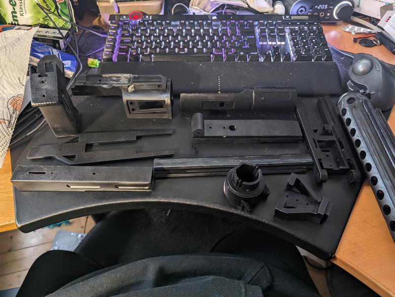 Around fifteen black resin parts laid out on a desk mat, including stock halves, receiver, barrel sections, rails and brackets