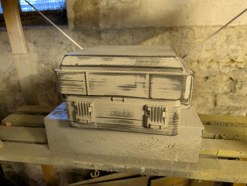Two instrument cases stacked on a concrete block in a cellar, painted in a bone base coat with black drybrushed edges and panel lines