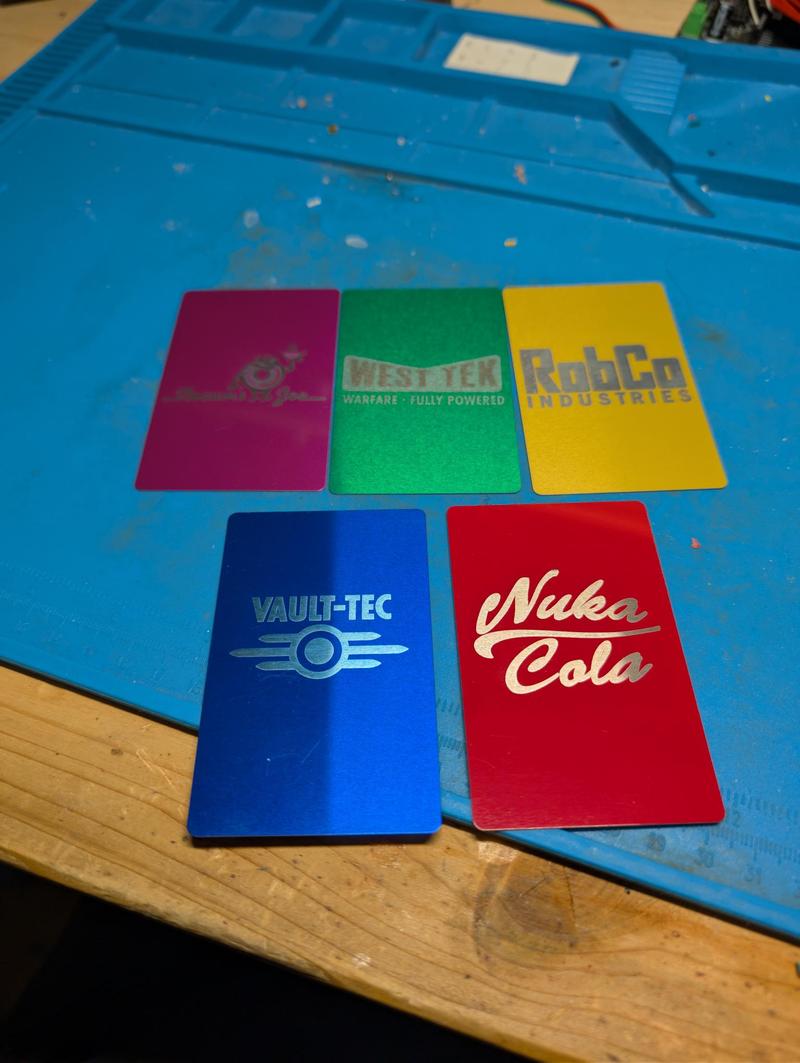 Five anodised aluminium cards in magenta, green, yellow, blue and red on a blue silicone mat, each laser-engraved with a different fictional corporate logo