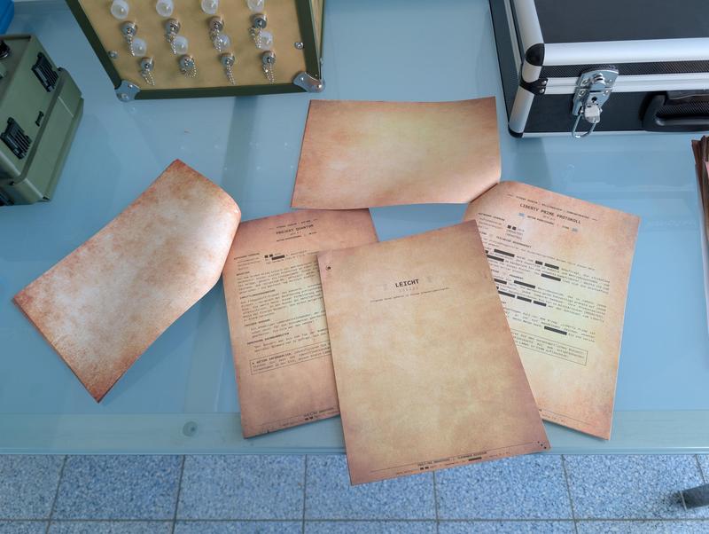 Five sheets of aged paper with typewriter-style printed briefing documents spread on a glass table next to an olive puzzle case