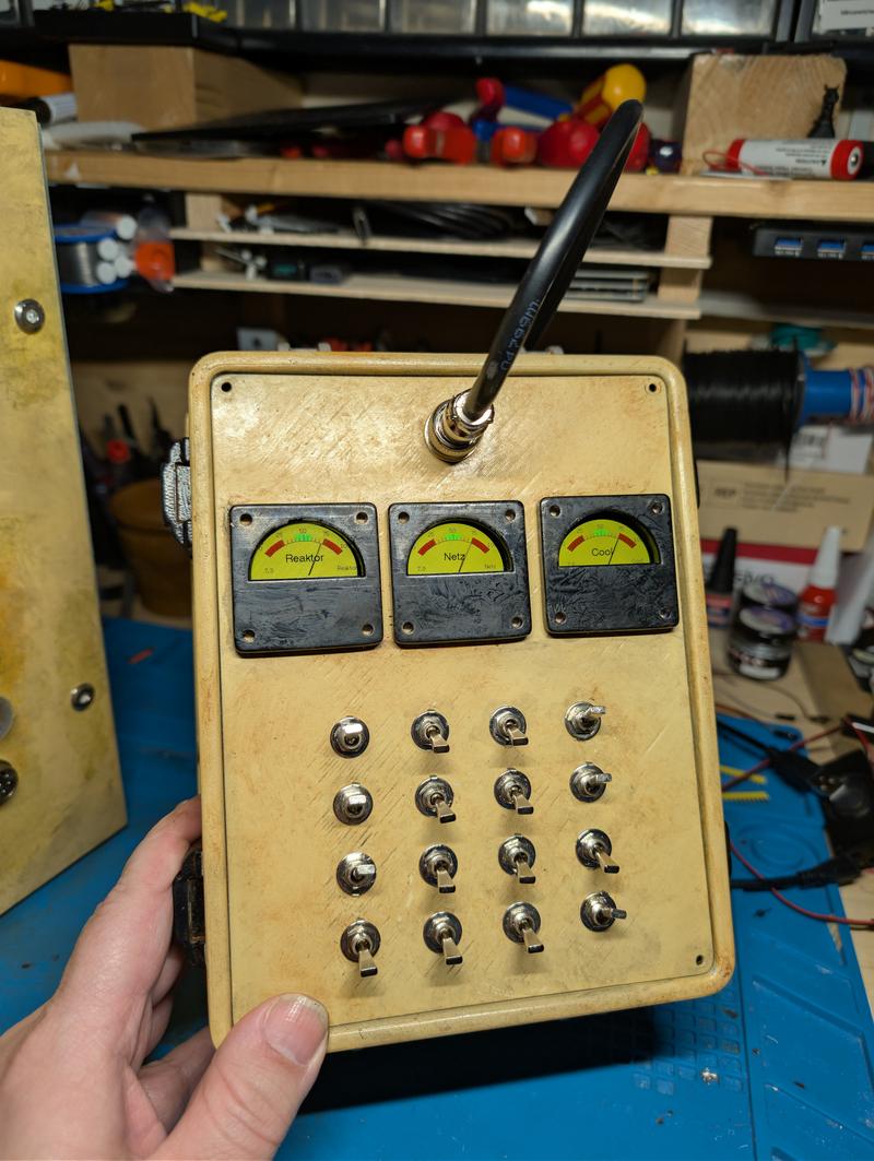 Bone-coloured weathered case held in one hand, with three simulated analogue meters labelled Reaktor, Netz and Cool above a four-by-four grid of chrome toggle switches