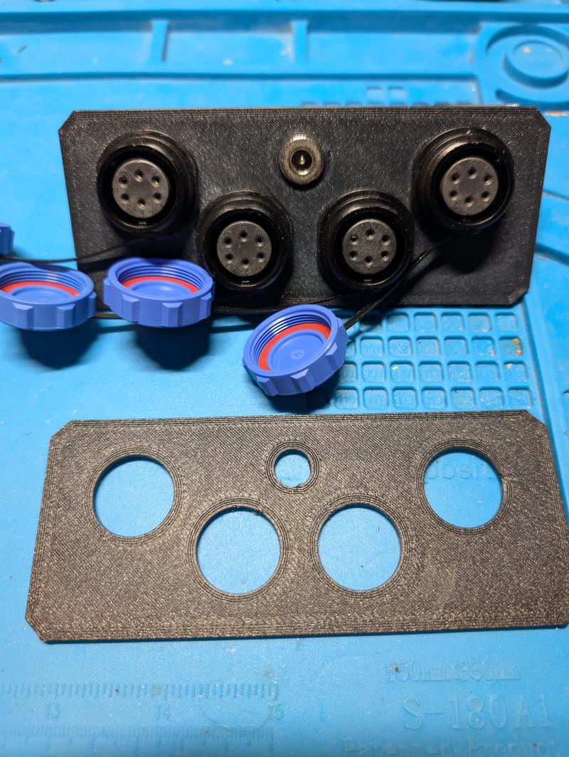 Black printed panel with four six-pin circular sockets and one smaller socket in the centre, with blue screw caps lying beside it and the matching cover plate below