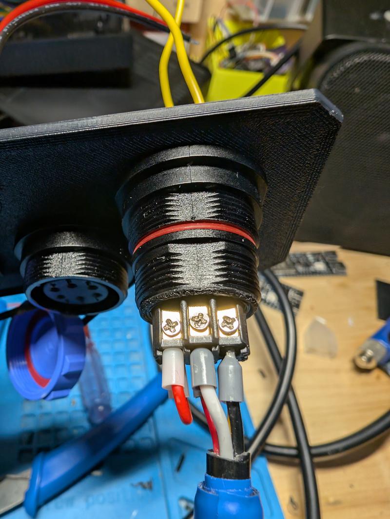 Close-up of the rear of a circular panel socket showing three screw terminals with ferruled wires, mounted through a printed panel