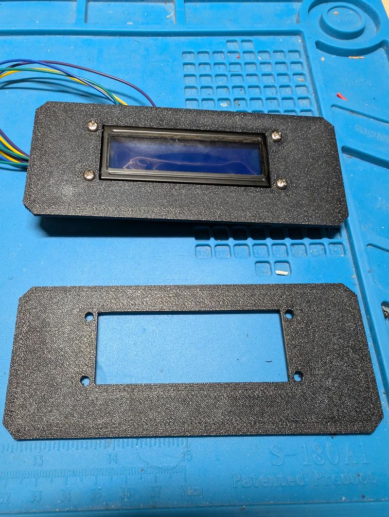 A 16 by 2 character LCD in a printed black bezel next to the matching cover frame on a blue mat