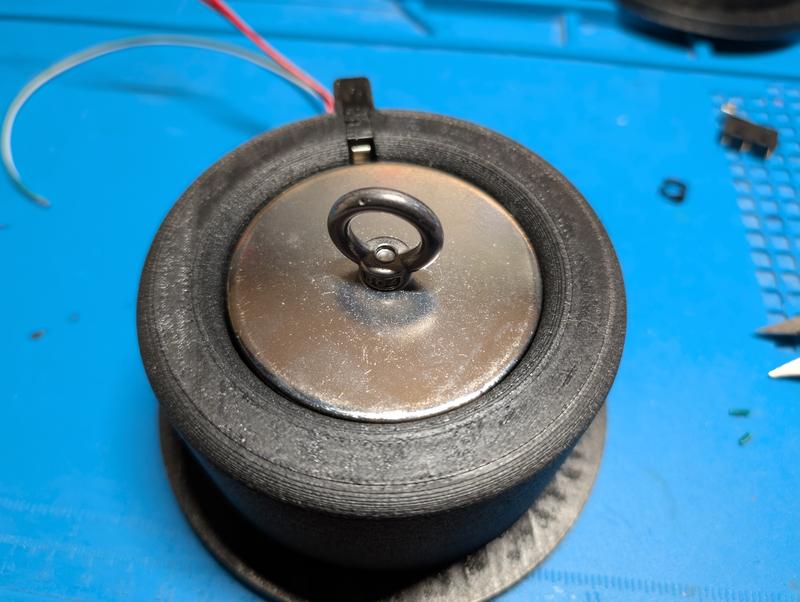 The same housing with a polished steel anchor plate resting on the magnet face, an eye bolt screwed into the centre of the plate