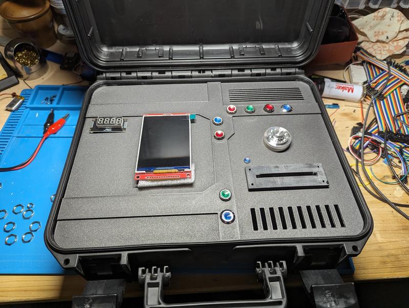 Unpainted grey printed panel resting in an open black transport case during dry fitting, with a TFT module, a four-digit display, illuminated buttons, a polished metal disc and the card slot placed in their cutouts