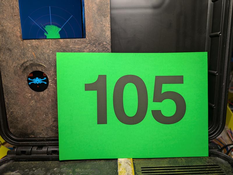 Console lid showing a four-inch screen with a blue radar sweep and green returns, a small round display below it with an animated blue X-wing and orange numerals, and a green test card marked 105 standing in front of the sensor