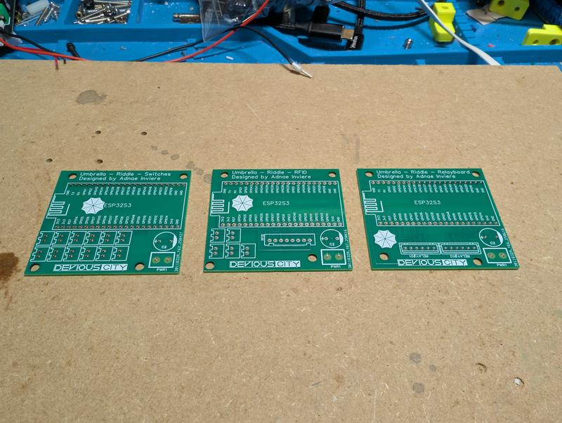 Three unpopulated green circuit boards laid side by side, labelled Umbrella Riddle Switches, Umbrella Riddle RFID and Umbrella Riddle Relayboard, each with an ESP32-S3 footprint and the Devious City logo