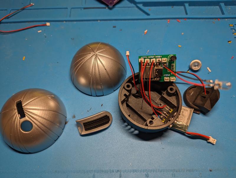 Disassembled detonator on a blue mat: two silver painted hemispheres, a printed switch cover, the open base with a green circuit board, wired LEDs, a lithium polymer pouch cell and a clear lens