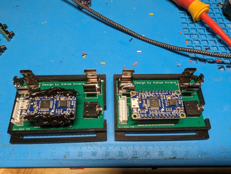 Two green carrier boards side by side in printed frames, each with metal battery clips and an Adafruit audio board with a micro USB port and stereo jack; the left board's audio module is covered in black potting compound