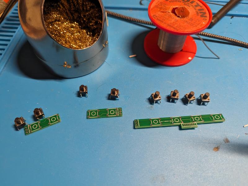 Loose tactile switches next to unpopulated green trigger boards on a blue mat, beside a solder spool and a brass tip cleaner