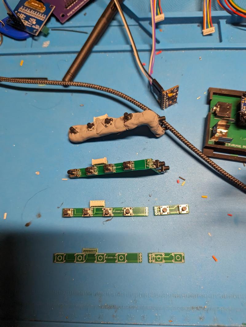 Several small green trigger boards on a blue mat in different stages: a bare four-position board, a populated one with tactile switches, and one fully encased in grey cured foam with a braided cable leaving it